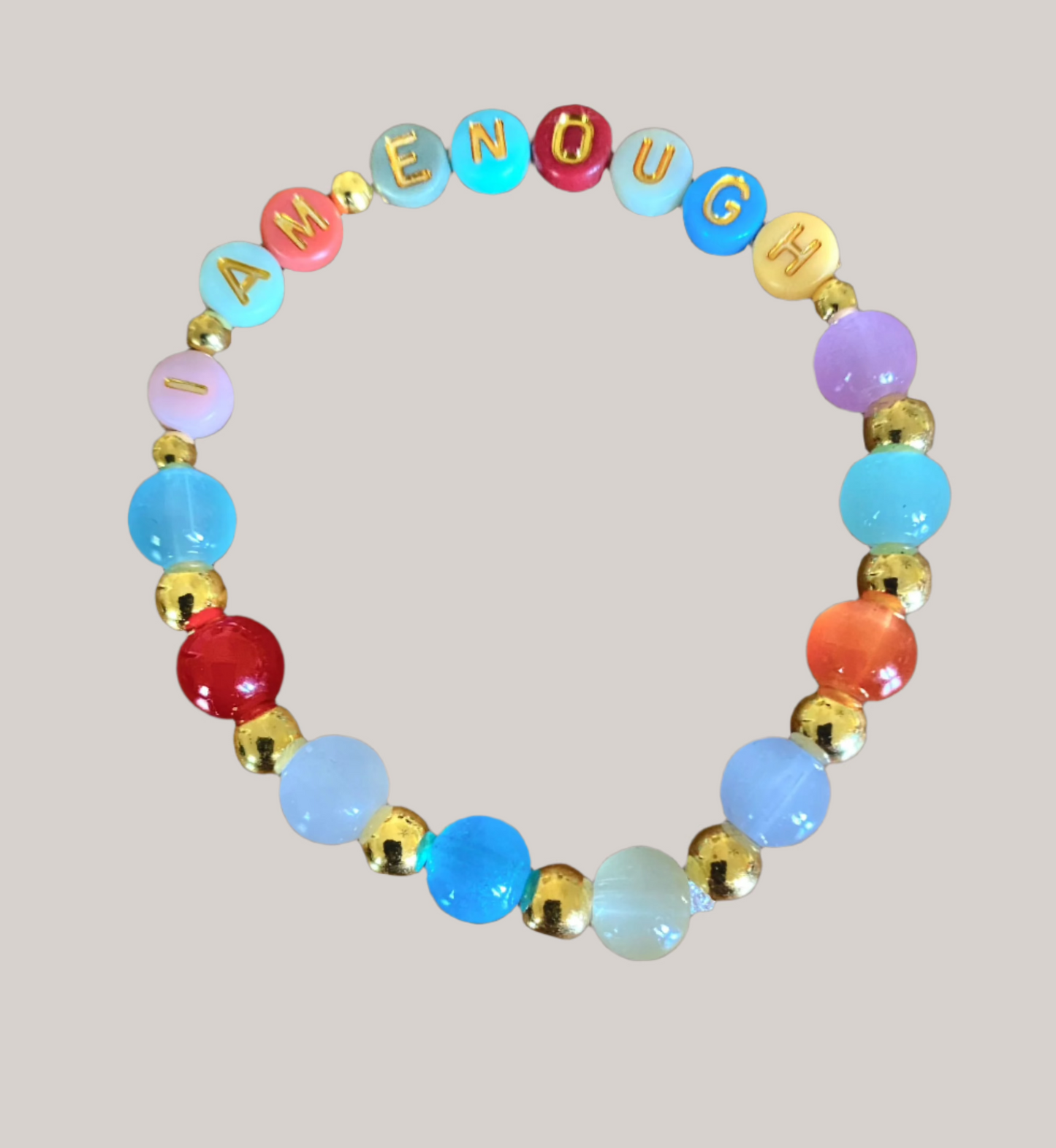 The I Am Enough Mercy Message Stretch Bracelet Set by Mercy Plus Grace features a vibrant, circular bracelet with alternating red, blue, purple, white, and gold glass beads. The words "I AM ENOUGH" are spelled out in gold letters on yellow beads, all elegantly displayed against a black background.