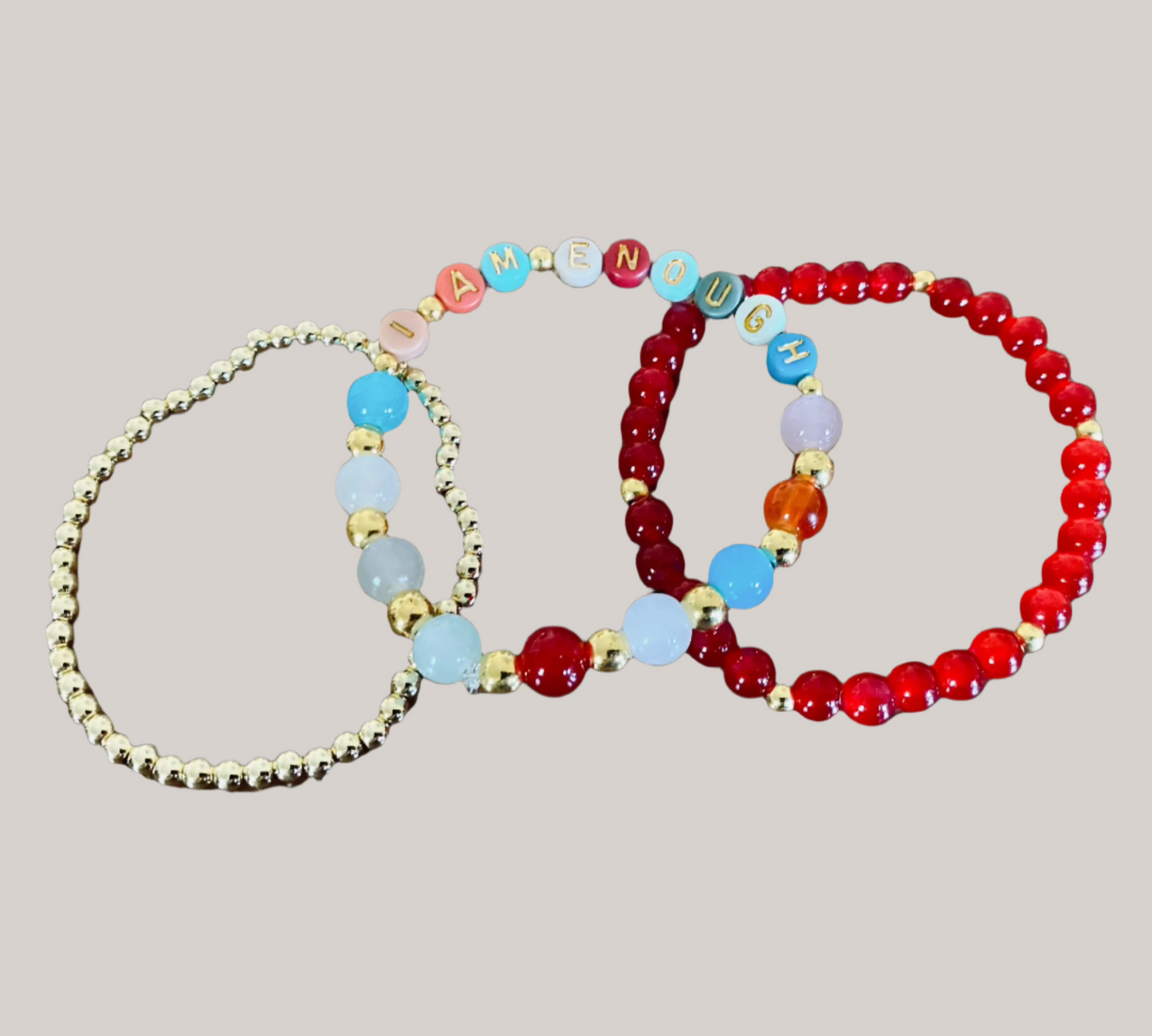 The I Am Enough Mercy Message Stretch Bracelet Set from Mercy Plus Grace features three stretch bracelets: a gold beaded one, a vibrant bracelet with letters spelling "I AM ENOUGH," and a red beaded bracelet adorned with colorful glass beads, including one orange bead and a few light blue beads, all set against a black background.