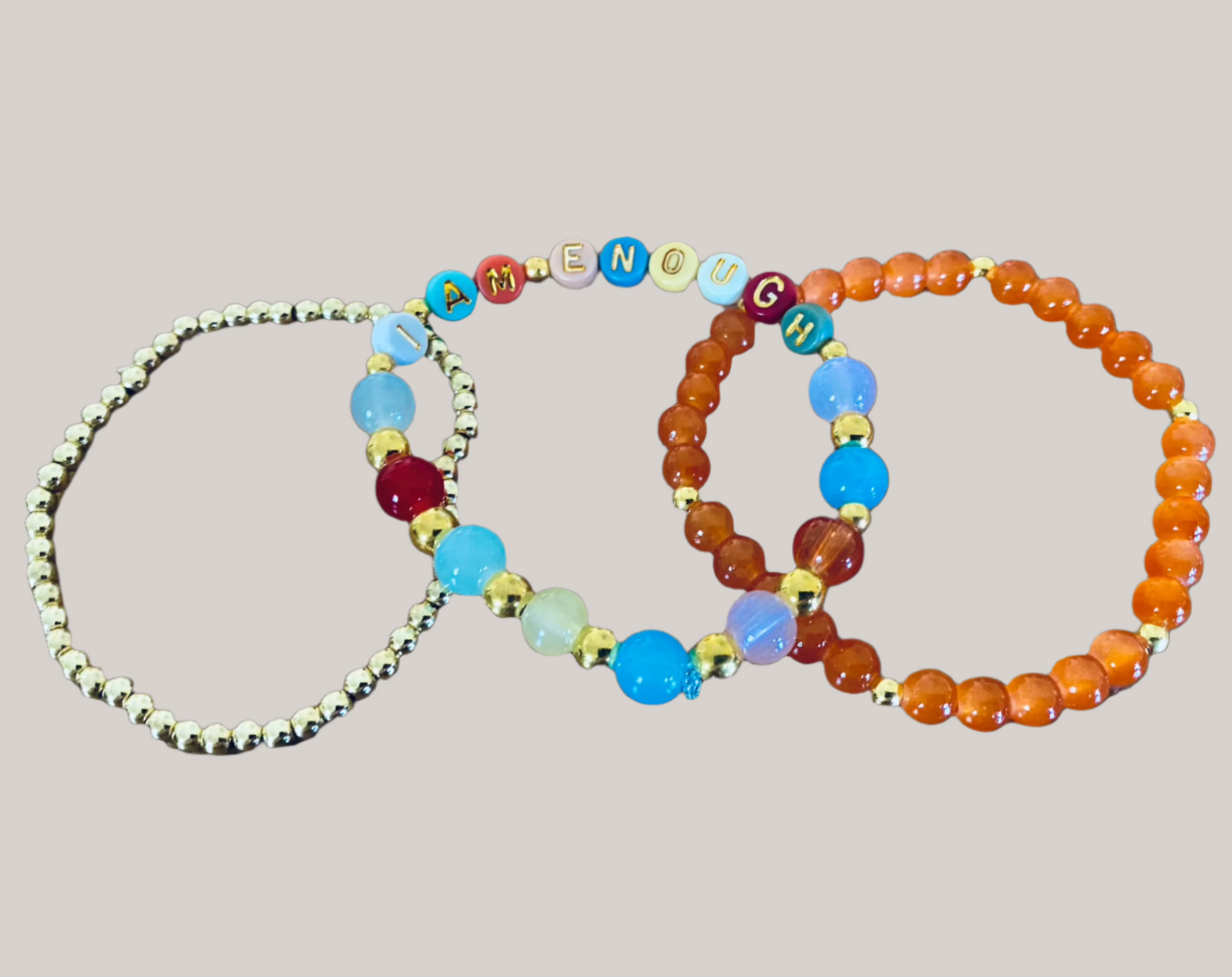 Three stretch bracelets from the Mercy Plus Grace "I Am Enough Mercy Message Stretch Bracelet Set" are displayed against a black background. The first bracelet is adorned with gold beads, the second features colorful blue, red, and white glass beads alongside multicolored letter beads that spell out "I AM ENOUGH," and the third bracelet consists of vibrant orange beads.